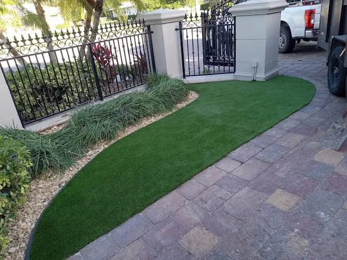 Curved artificial turf bordering a beautiful paver driveway near an ornate metal gate